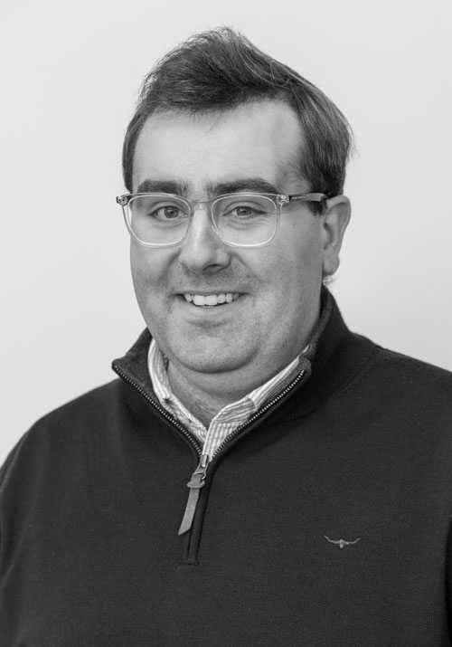 craig-bebbington - Sellar & Sellar Portrait of a smiling man wearing rectangular glasses and a dark zip-up sweater over a collared shirt.